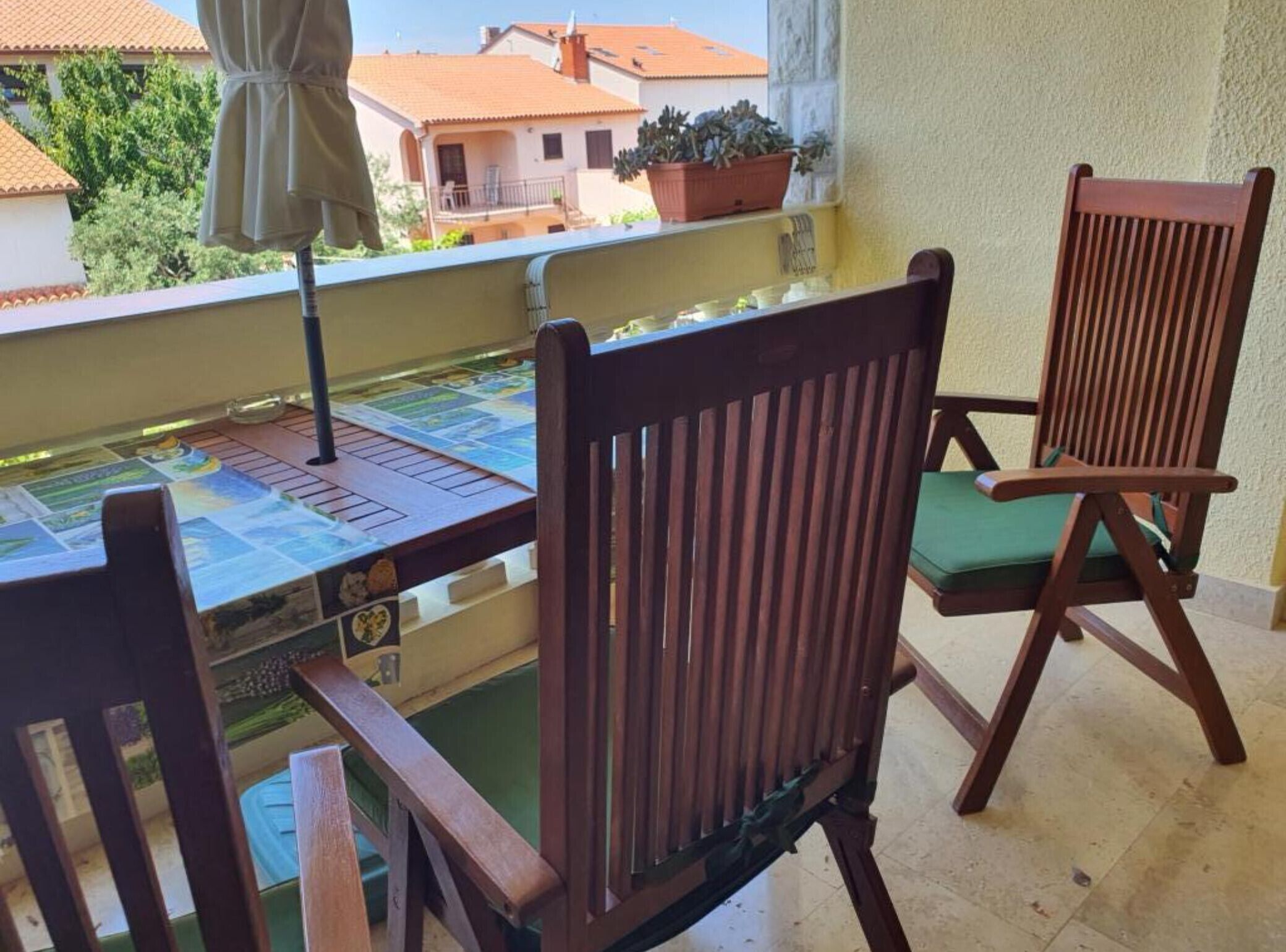 A balcony with two wooden chairs, one with a green cushion, and a table with a colorful tablecloth.