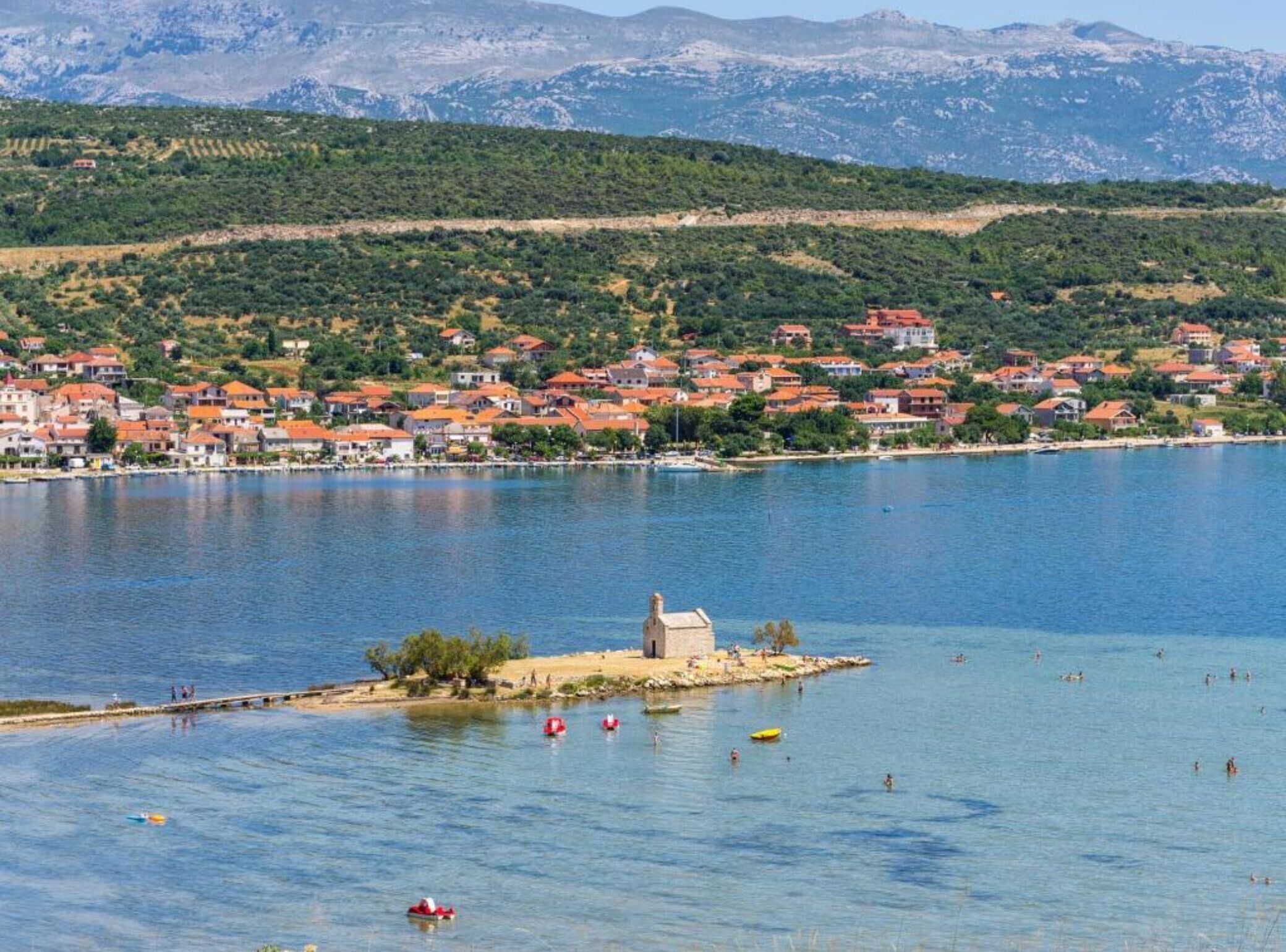 Un pueblo costero con casas a lo largo de la orilla, una pequeña isla con un edificio, y montañas al fondo.