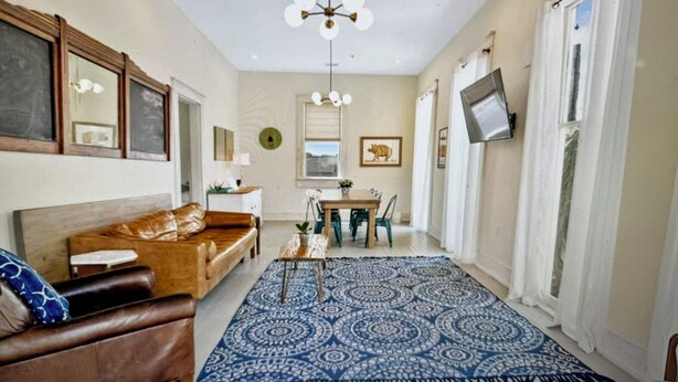A living room with a brown leather sofa, a blue patterned rug, a dining table with chairs, a chandelier, and a television mounted on the wall. - Beautiful upstairs apartment for rent monthly near Audubon park (New Orleans)