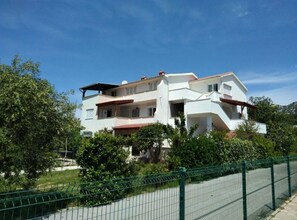A two-story white building with a red-tiled roof, surrounded by greenery and a green fence. - Apartments Verović Rab Banjol - Two Bedroom Apartment With Terrace (A1) (Banjol)