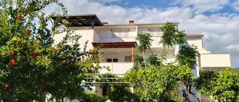 A two-story house with a balcony, surrounded by greenery and a clear sky.
