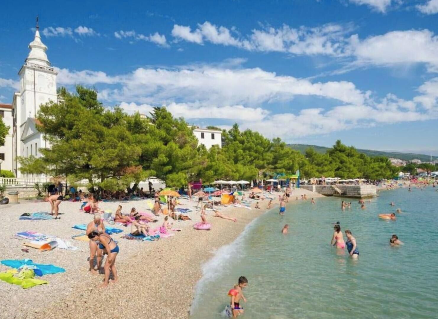 A beach with people sunbathing and swimming, a white building with a tower, and trees in the background.