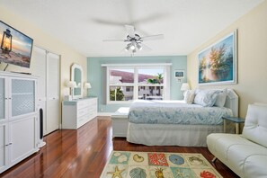 Una camera da letto con un letto, un comodino, una cassettiera, una ventola a soffitto, una finestra vista edifici e un tappeto a tema spiaggia.