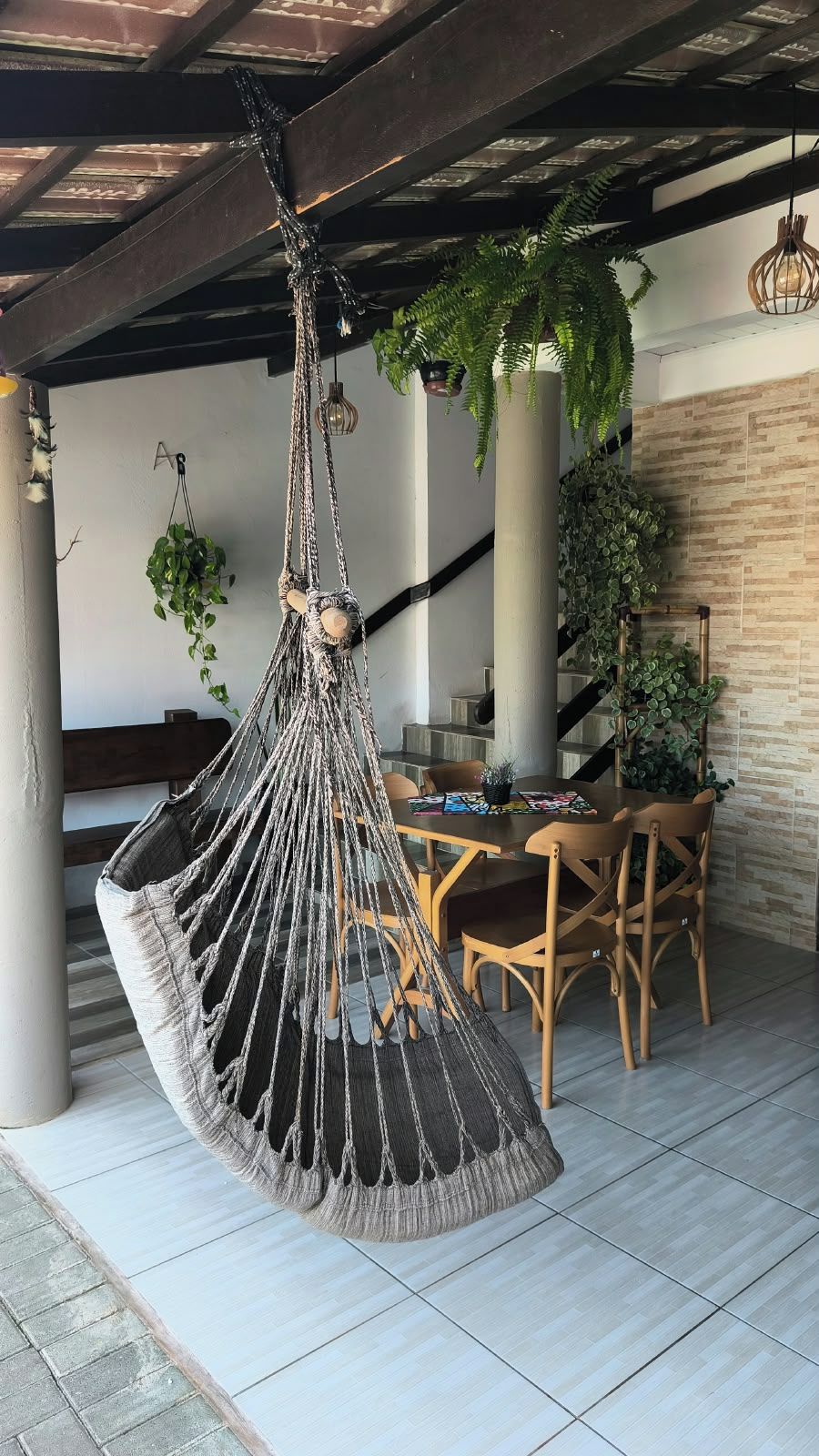A hanging macrame chair in a covered outdoor area with a dining table and chairs, surrounded by potted plants.