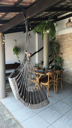 A hanging macrame chair in a covered outdoor area with a dining table and chairs, surrounded by potted plants. - Pousada Cantinho da Jake (Penha)