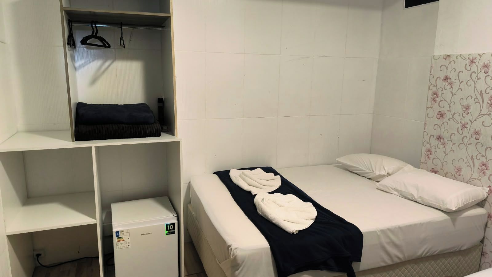 A single bed with white bedding and a black blanket, a small bedside table, a wardrobe with hangers, and a white wall with a floral pattern.