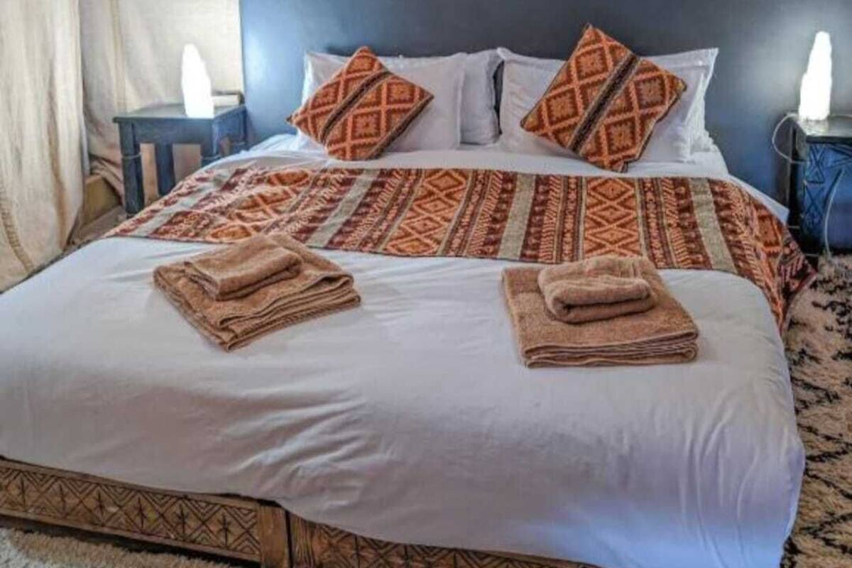 Una cama con almohadas estampadas, una toalla doblada, un cabecero y una mesita de noche con una lámpara.