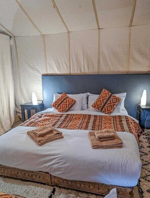 A bed with patterned pillows and a folded towel, a headboard, and a nightstand with a lamp. - Unique Sahara Camp (Merzouga)