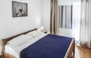 Un lit bien fait, avec une literie blanche et bleue, une petite table de chevet avec une lampe, et un tableau floral encadré accroché au mur.