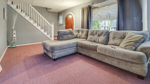 Ein Wohnzimmer mit einem grauen Ecksofa, einer Treppe mit weißen Geländern und einem großen Fenster mit Vorhängen.