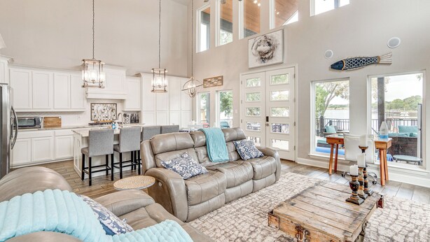 A spacious living area with a leather sofa, a coffee table, and a dining table with chairs. There is a kitchen area with white cabinets and a refrigerator. Large windows provide natural light and views of the outdoors. - Private Dock & Yard! Modern Getaway in Mabank (Mabank)