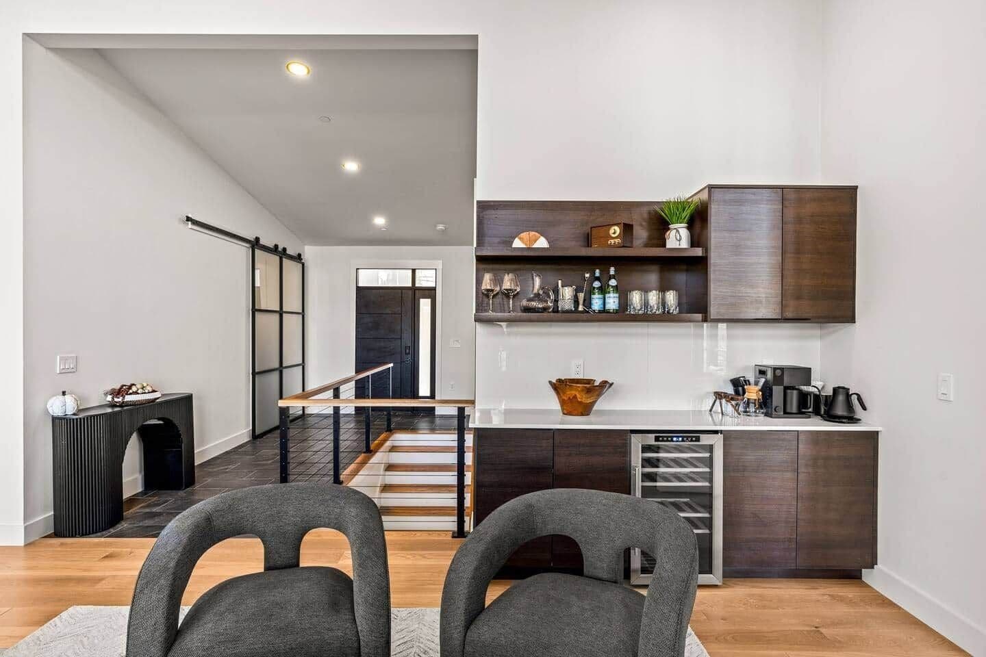 A modern kitchen with dark wood cabinets, a built-in oven, and a coffee machine. There is a staircase leading to another level, and a dining area with two grey chairs.