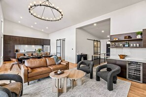 A spacious living area with a brown leather sofa, grey armchairs, and a wooden coffee table. A modern kitchen with dark cabinetry and a dining area with a table set for four is visible. - Luxury Mountain Estate, Sleeps 12, Theater, Games & Hot Tub (Campton)
