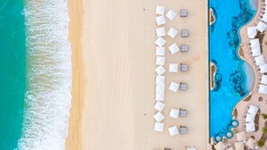 Een luchtbeeld van een strand met witte parasols en ligbedden langs de kustlijn, een helderblauw zwembad grenzend aan het strand, en een zandstrand.