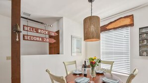 Un coin repas avec une table en bois dressée pour quatre personnes, des chaises blanches, un luminaire suspendu et un panneau mural indiquant les directions.