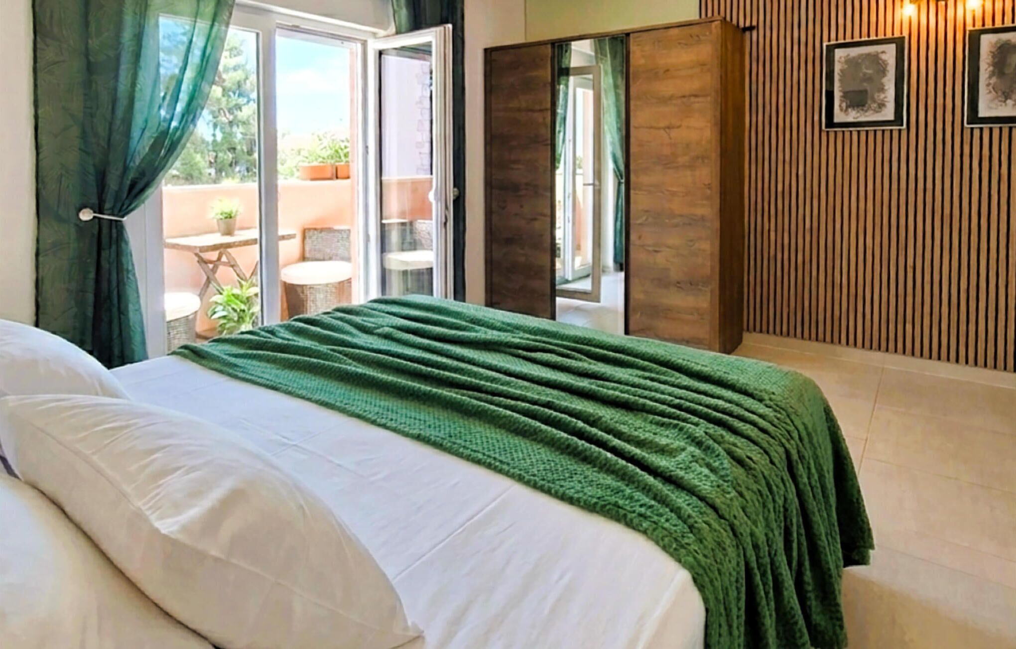 A bedroom with a bed, a green blanket, a sliding door to a balcony, and a wooden wall with framed pictures.