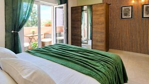 Un dormitorio con cama, manta verde, puerta corredera a un balcón y pared de madera con cuadros.