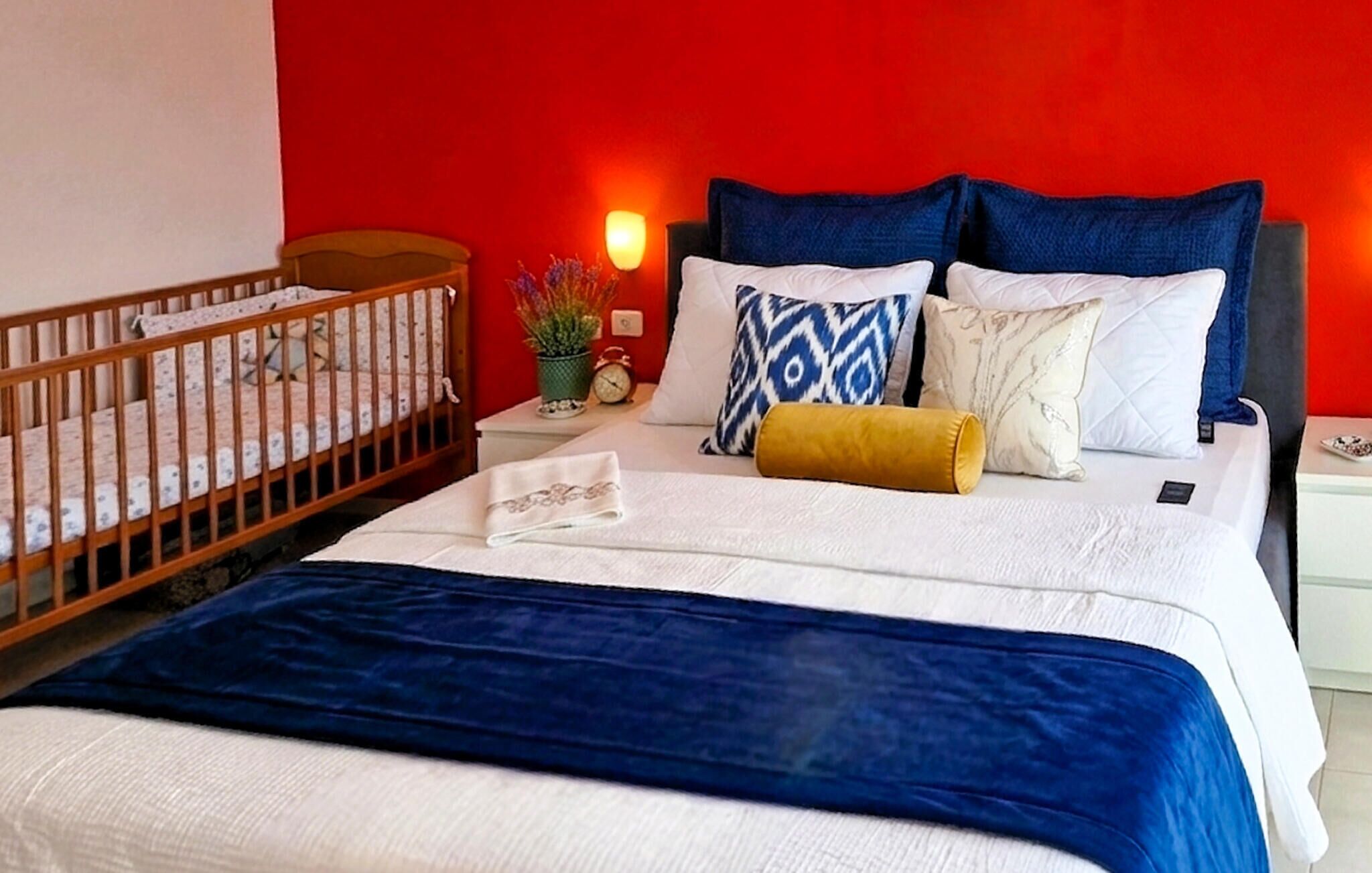 Une chambre d’hôtel avec un berceau, un lit recouvert d’une courtepointe bleue et une table de chevet avec une lampe et une horloge.