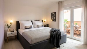 Ein modernes Schlafzimmer mit einem großen Bett, Nachttischen, einem Beistelltisch, einem Fenster mit Blick und einem Balkon mit Gartenmöbeln.