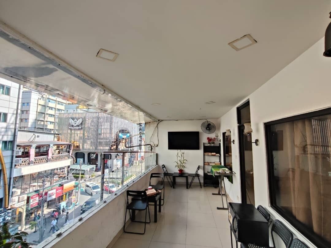 A balcony with a view of a busy street, a table with chairs, a fan, and a TV mounted on the wall.