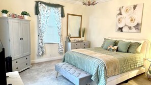 Een slaapkamer met een bed, nachtkastje, dressoir, bankje, spiegel en schilderij.