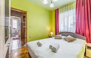 A hotel room with a bed, a nightstand, a lamp, and a view of the outside through a window with red curtains. - Beautiful apartment in Pula with outdoor swimming pool (Pula)