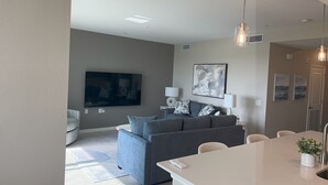 Un salon moderne avec un canapé gris, un téléviseur à écran plat et un coin repas comprenant une table et des chaises blanches.