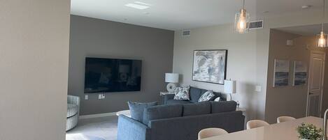 Ein modernes Wohnzimmer mit grauem Sofa, einem Flachbildfernseher und einem Essbereich mit weißem Tisch und Stühlen.