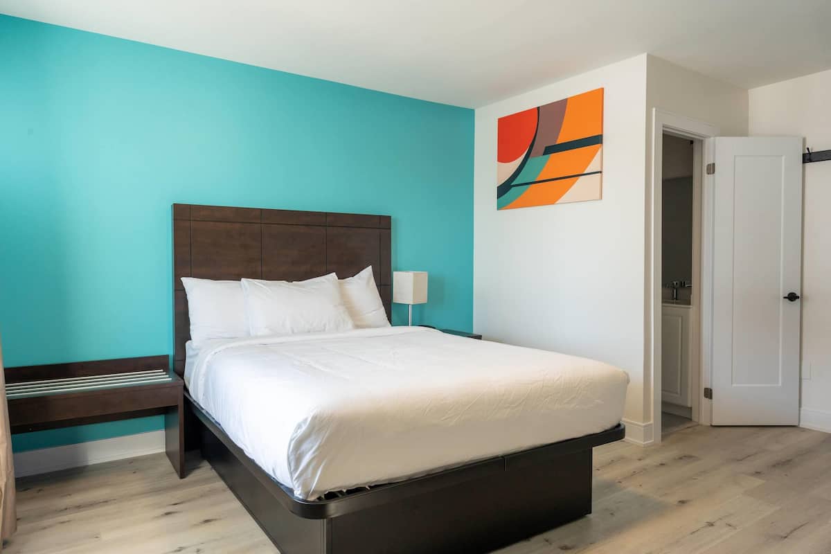 A bedroom with a bed, a nightstand, a wall-mounted lamp, and a door leading to a bathroom.