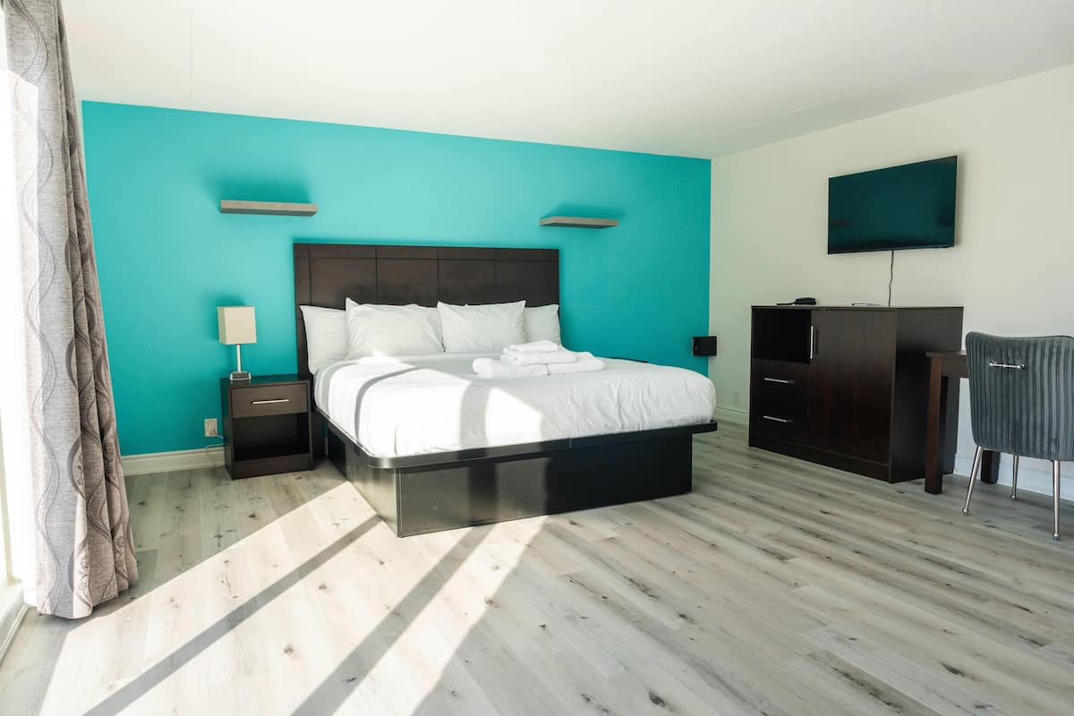 A modern hotel room with a large bed, a flat-screen TV, a dark wooden wardrobe, and a small desk with a lamp.