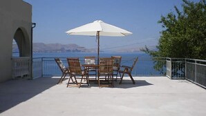 Un patio con una mesa y sillas bajo una sombrilla blanca, con vista a una masa de agua y terreno distante.
