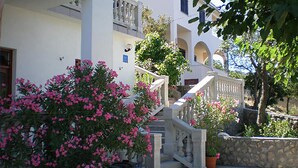 Un edificio blanco con una escalera que lleva a un nivel superior. Hay plantas con flores rosas en primer plano.