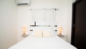 Cama bem arrumada com roupa de cama branca, duas luminárias de cabeceira, um encosto de cabeça branco e uma cortina branca.