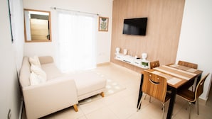 Uma sala de estar moderna com um sofá, uma mesa de jantar com cadeiras, uma TV fixada na parede e uma janela com cortinas.