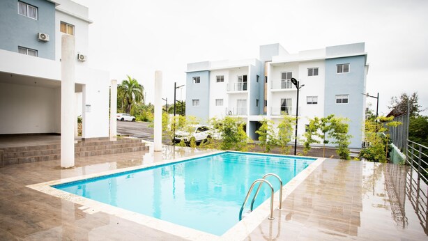 Uma piscina com deck ladrilhado, rodeada por edifícios modernos com varandas e um carro estacionado ao fundo.