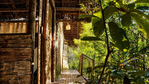 Un sendero de madera que conduce a una construcción rústica con techo de paja y una lámpara colgante.