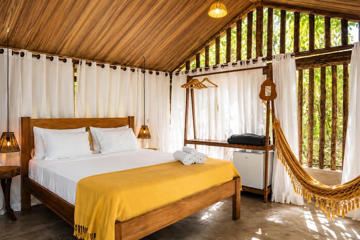 Una cama con estructura de madera, ropa de cama blanca, una manta amarilla, una mesita de noche con una lámpara y una hamaca.