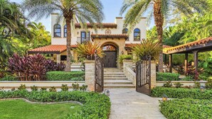 A Mediterranean-style villa with a central entrance, a stone staircase, and a well-maintained garden with palm trees and shrubs. - Mediterranean Waterfront Estate on Caloosahatchee (Fort Myers)