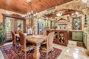 Un comedor amplio con una mesa y sillas de madera, una lámpara de araña y una cocina con armarios verdes y una pared de ladrillo.