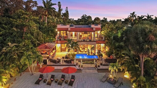 A two-story building with a swimming pool, surrounded by palm trees and a sandy beach. - Mediterranean Waterfront Estate on Caloosahatchee (Fort Myers)