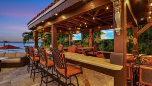 An outdoor bar area with a stone wall, wooden ceiling, and multiple bar stools. - Mediterranean Waterfront Estate on Caloosahatchee (Fort Myers)
