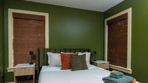 A neatly made bed with white linens, a green upholstered headboard, and multiple pillows in various colors. There are two bedside tables with lamps, and the room has green walls and wooden flooring. - Fenced Yard, Parking + Workspace - Downtown Dallas (Dallas)