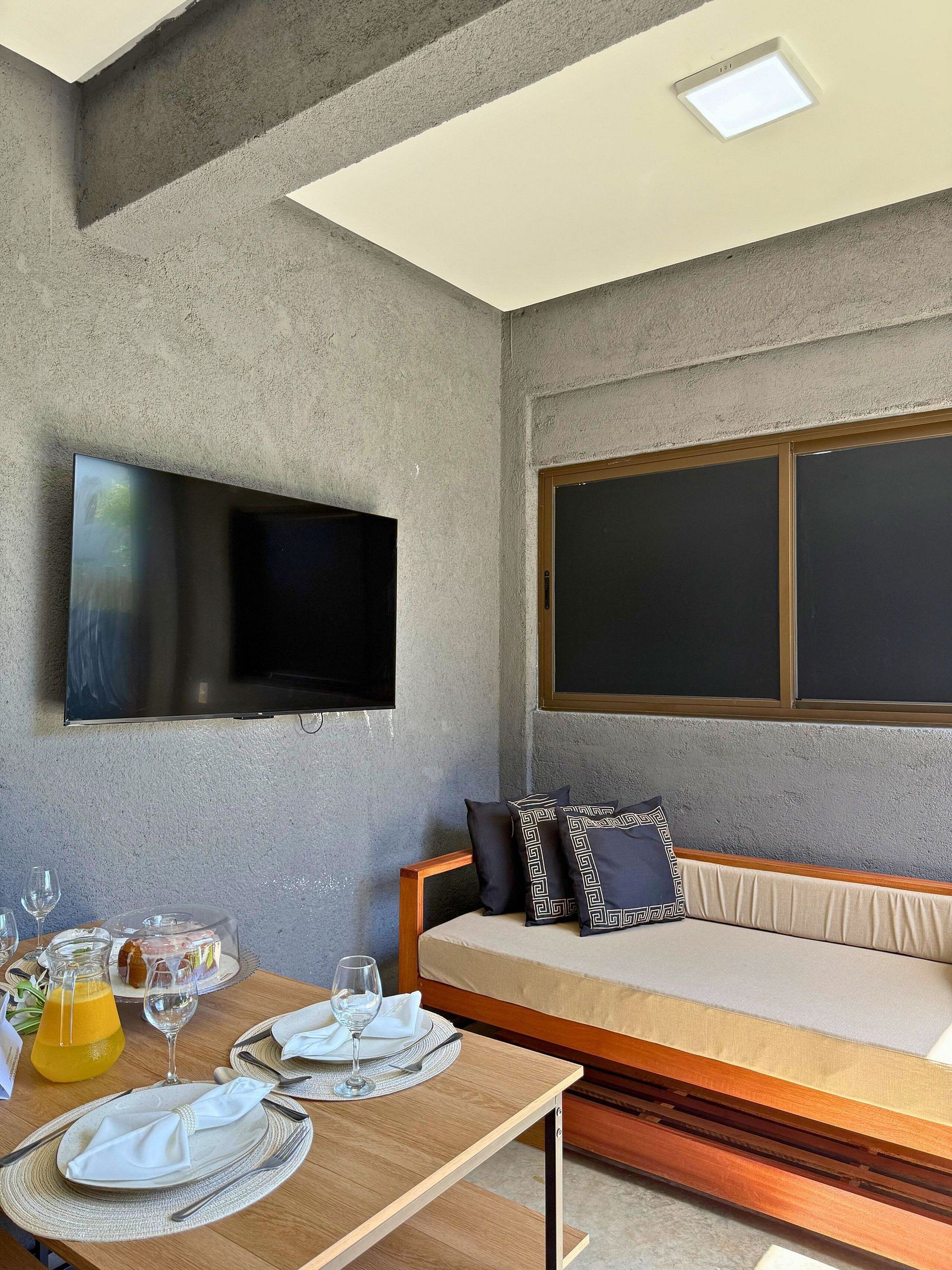 A modern room with a flat-screen TV, a sofa with patterned cushions, a dining table set for two, and a large window.