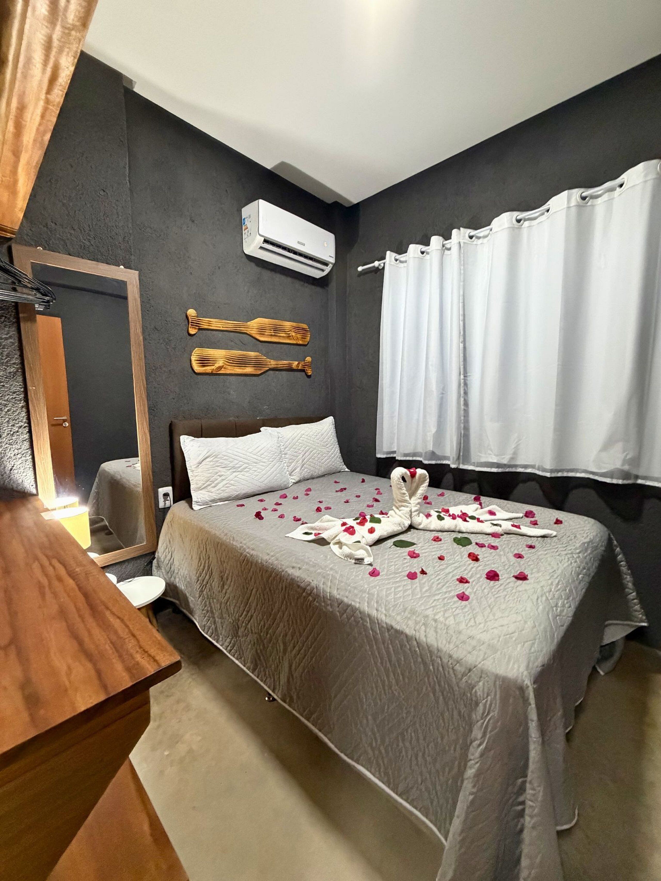 Een hotelkamer met een bed, een houten hoofdeinde, een witte airconditioning, een spiegel en een raam met witte gordijnen.