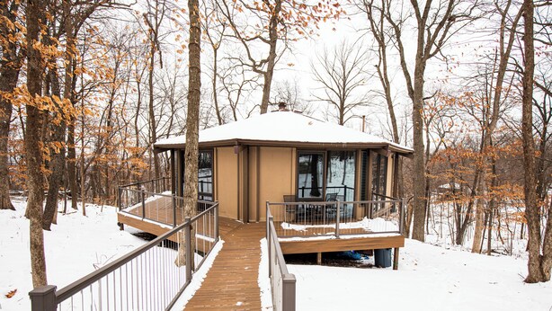Une terrasse en bois mène à un bâtiment entouré d’arbres, et il y a de la neige par endroits sur le sol.