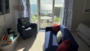 Een woonkamer met een bank, fauteuil en een televisie. Er is een balkon met een tafel en stoelen.