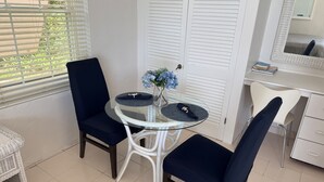 Een kamer met een ronde glazen tafel, twee donkerblauwe stoelen, een witte deur met spiegel en een raam met jaloezieën.