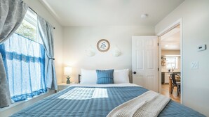 A bedroom with a large bed, a bedside table with a lamp, a wall clock, and a door leading to a kitchen.