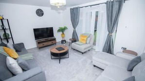 A modern living room with a sofa, armchair, and ottoman, a TV, a plant, and a clock on the wall.