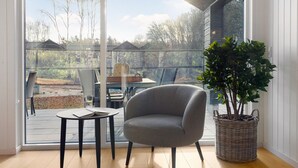 A modern room with a large sliding glass door leading to an outdoor patio. Inside, there is a grey armchair, a small table with a book, and a potted plant. - Cozy home in Glesborg with sauna (Glesborg)